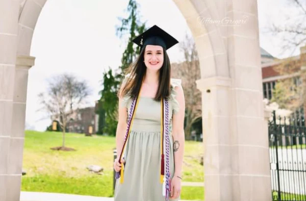 Photo of a smiling graduate woman in cap and gown, standing in front of the Marywood University archway, with a statue above the arch and a clear sky in the background.